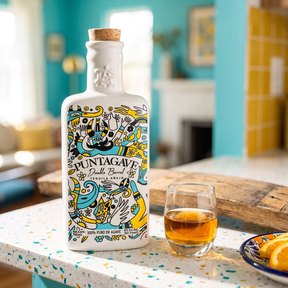 Puntagave Double Barrel Añejo Tequila, Ceramic Edition, 82 proof, on a terrazzo home bar with a copita of añejo, an oak barrel stave and orange slices on a Talavera dish