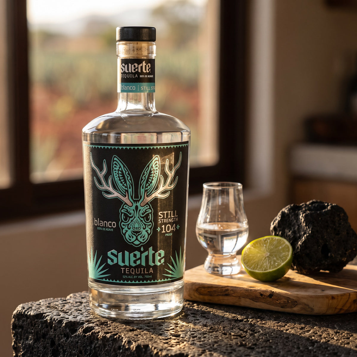 Suerte Still Strength Blanco Tequila, 104 proof, on a volcanic stone home bar with a copita of blanco and a halved lime, agave fields in the window
